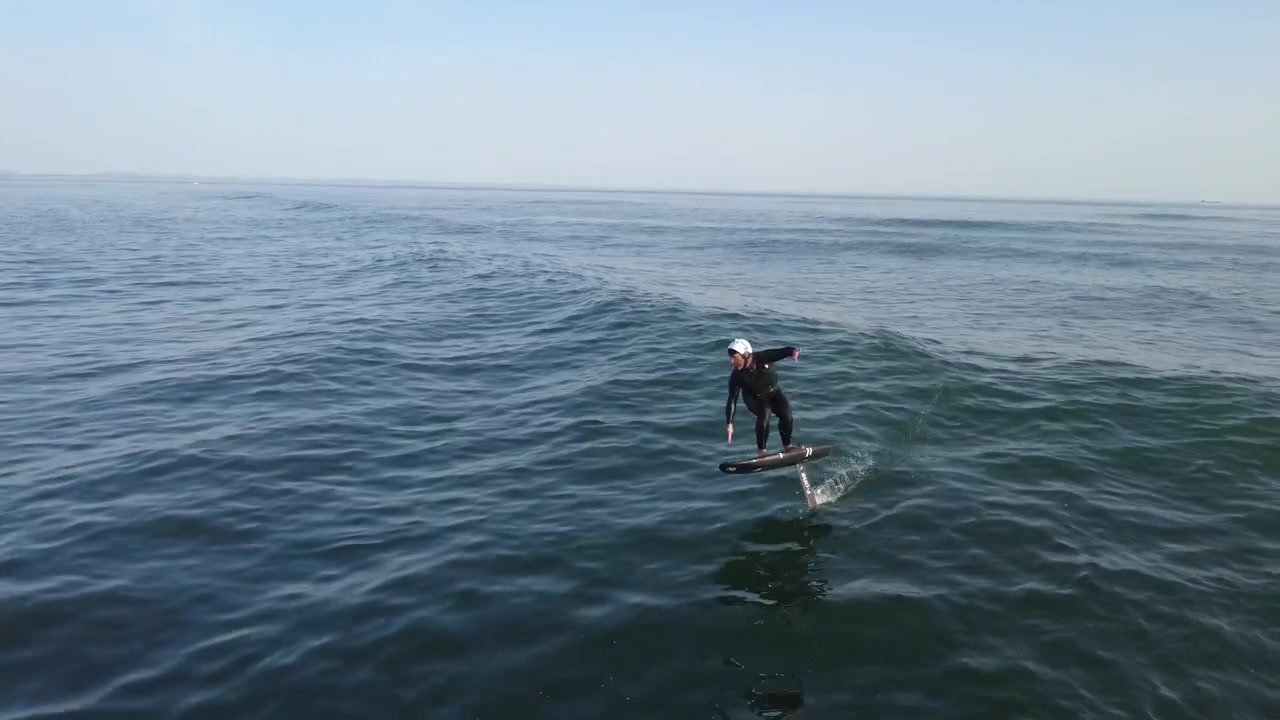 Guarda un rider di surf foil in posizione prona che pompa, esegue curve pulite e potenti roundhouse mentre cavalca le onde dell'oceano con velocità, fluidità, equilibrio e precisione