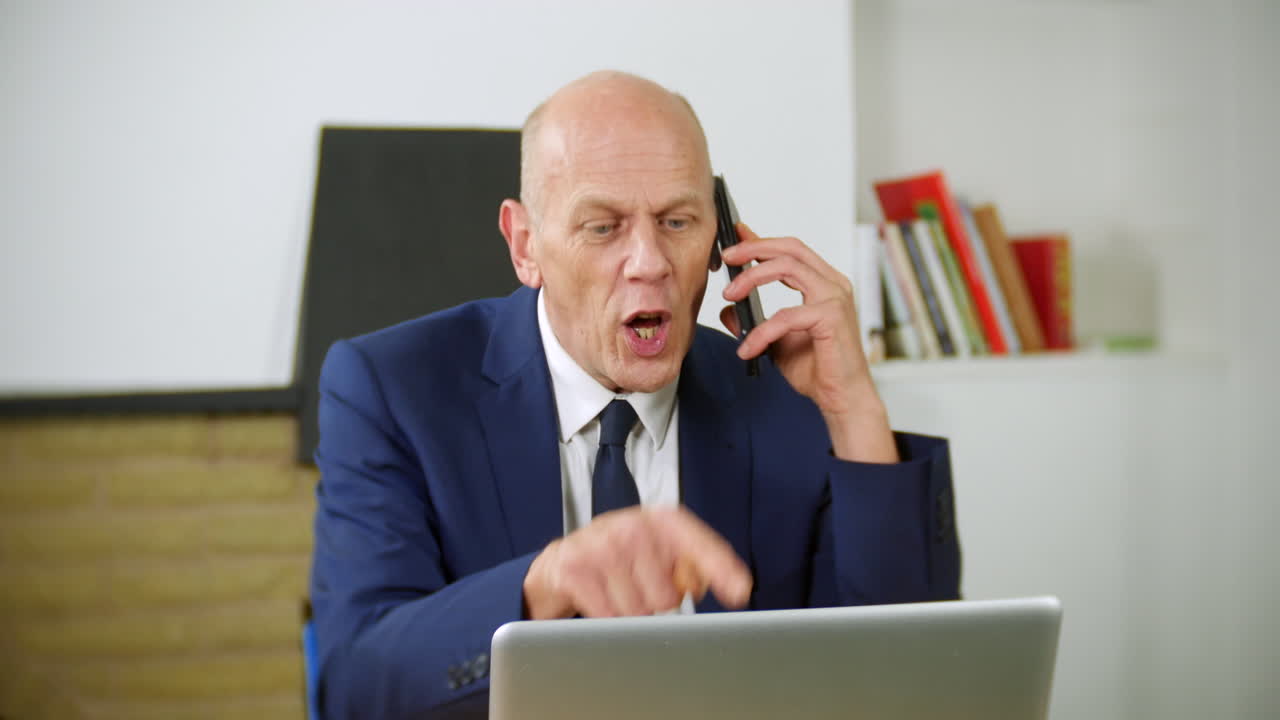 An angry mature businessman is talking on the phone in his home office