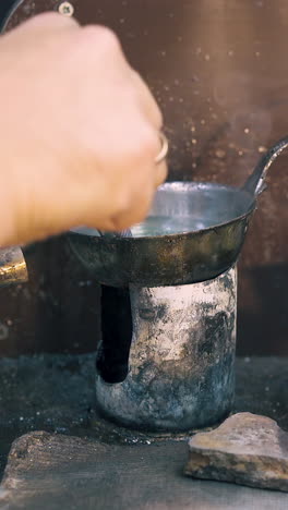 cu: jeweler takes off gold detail from metal bowl with boiling green liquid over gas burner on table close view