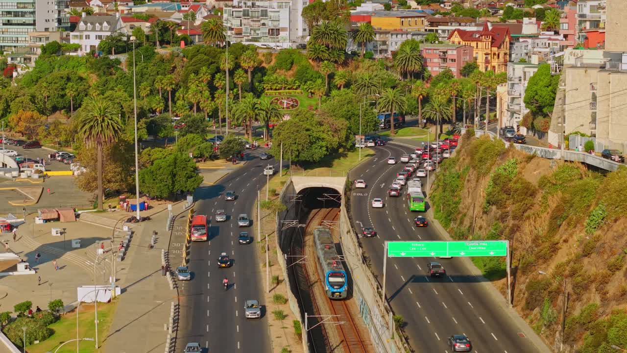 Luftaufnahme der Metrostrecke Valparaíso-Limache mit einem einfahrenden Zug in einen unterirdischen Tunnel, mit der Blumenuhr von Viña del Mar auf dem Castillo-Hügel, Chile.