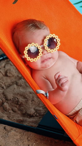 newborn caucasian boy in shorts lies in baby chair. little kid in sunglasses on the imitated beach. vertical video