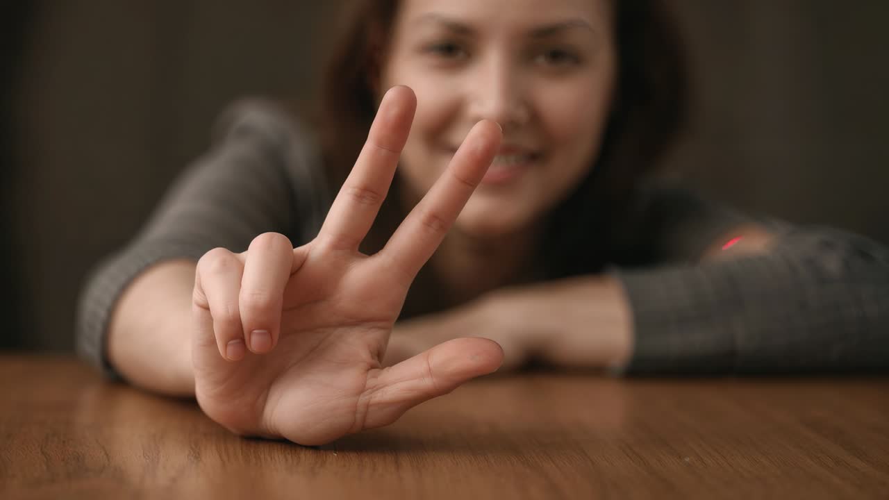 Camera focus shifting, leaning woman in grey sweater on wood table showing V sign greeting camera