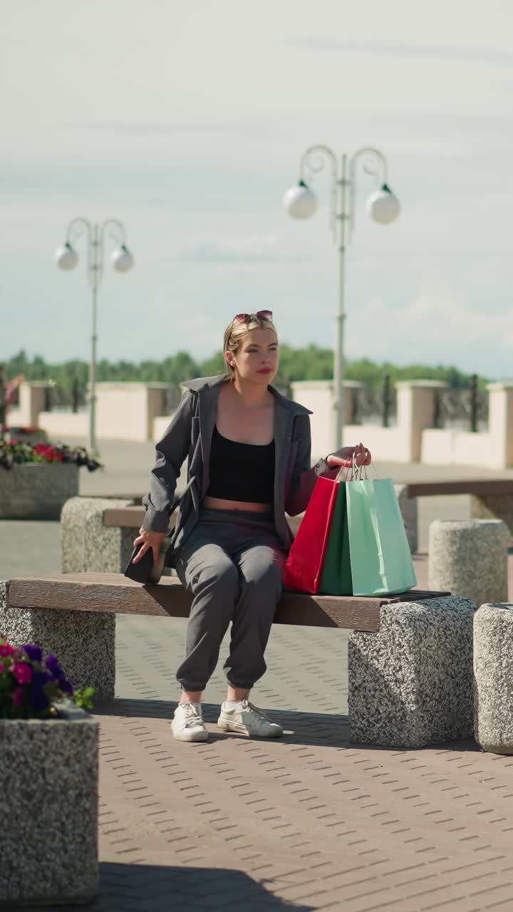 dama con tres bolsas de compras camina hacia bancos al aire libre, coloca las bolsas en el banco, y ajusta su bolso negro mientras está sentada, el fondo incluye flores y bancos