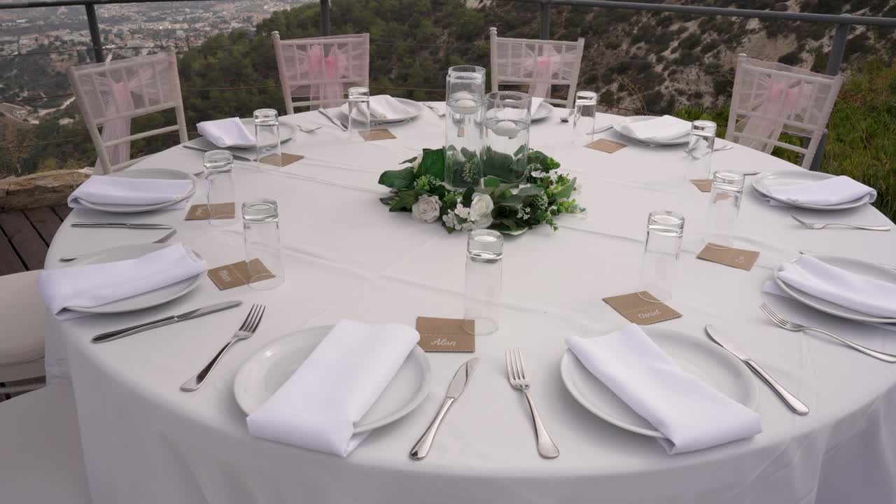 An elegant round table is beautifully arranged for a special event, featuring fresh flowers and tableware. The scenic background adds a touch of tranquility to the setting