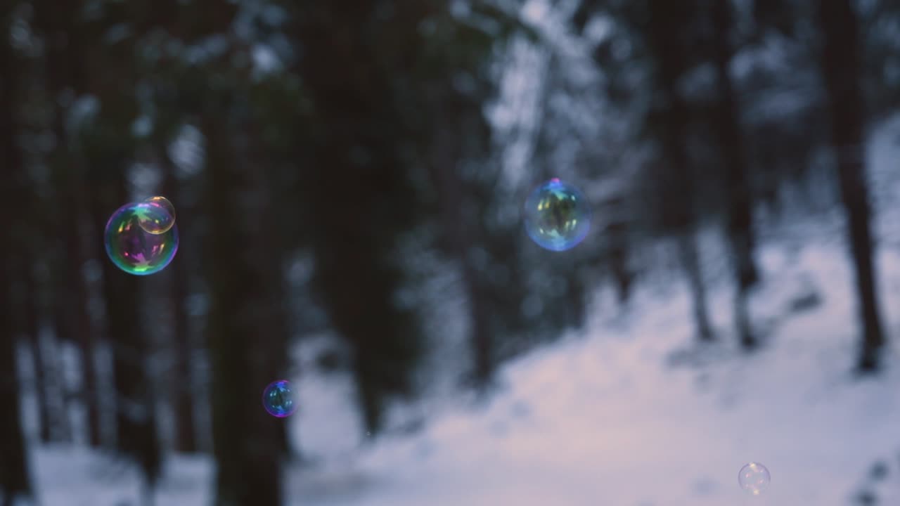 Sateenkaarenväriset saippuakuplat leijuvat ilmassa, taustalla lumimetsä.
