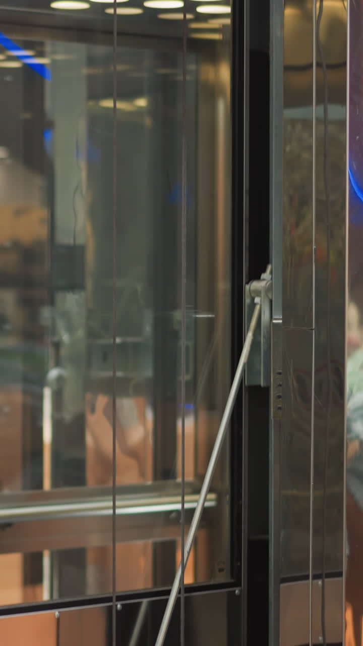 쇼핑몰에서 움직이는 현대적인 유리 엘리베이터, 우아한 반사, 기계적 세부 사항, 그리고 주변 인테리어와 광고에서 흐릿한 생생한 색을 보여줍니다.