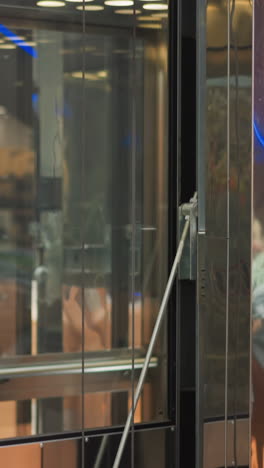 un moderno ascensor de vidrio que se mueve en un centro comercial, mostrando reflejos elegantes, detalles mecánicos y colores vibrantes borrosos de los interiores y anuncios circundantes