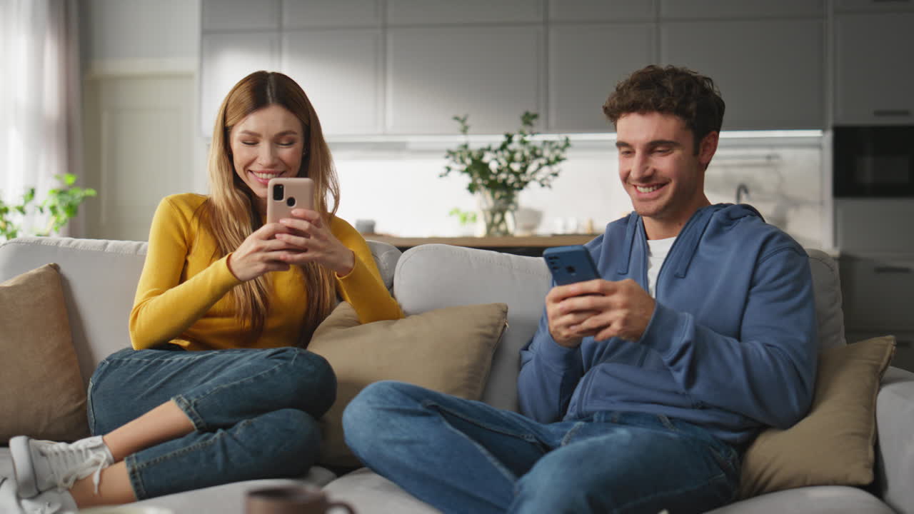 Happy newlyweds chatting smartphones at domestic weekend. Couple relaxing sofa