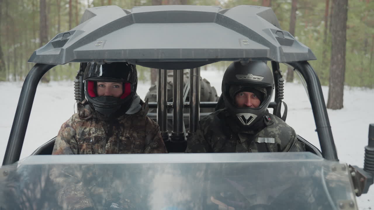 Vista frontale di un duo in ATV con caschi. Due piloti mimetizzati siedono fianco a fianco all'interno di un UTV su un sentiero di pini innevato. Il conducente afferra l'acceleratore mentre il passeggero scruta il percorso. Sono visibili un auricolare radio e giacche protettive.