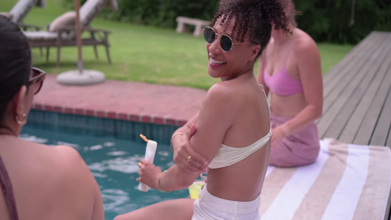 Female friends lounging at private pool, sun warming skin, woman in white bikini applying sunscreen