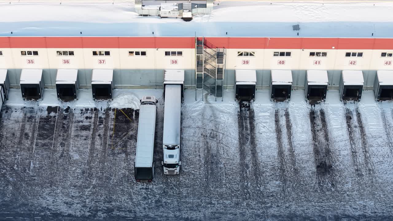 Dronevideo af en sneklædt industriel læsserampe med lastbiler på række, der fremhæver effektiv logistik i en kold og vinterlig omgivelse. Overheadperspektivet understreger storheden.