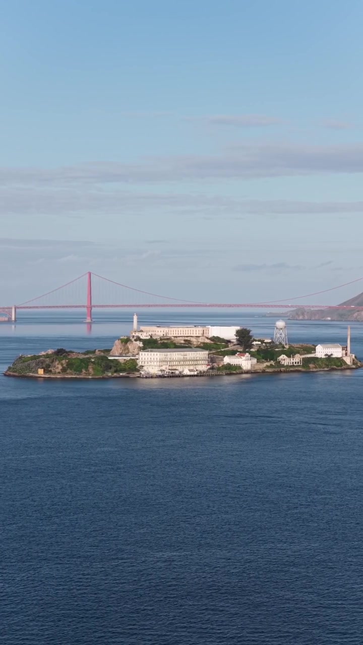 O complexo prisional da Ilha de Alcatraz fica na Baía de São Francisco, com as torres da Ponte Golden Gate estendendo-se sobre a água ao fundo, a luz do pôr do sol lançando um brilho quente sobre a costa da Califórnia. Imagem aérea vertical ampla de drone.