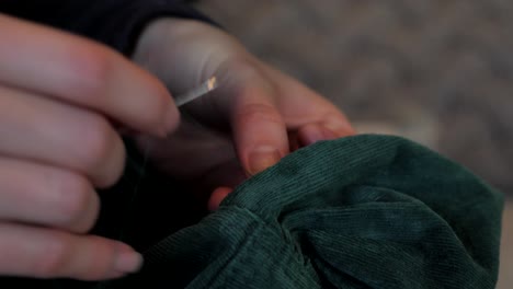 female hands sew green jacket with thin metal needle and string thread to make alteration and tailoring adjustment, side perspective macro view as stitching needle goes through material