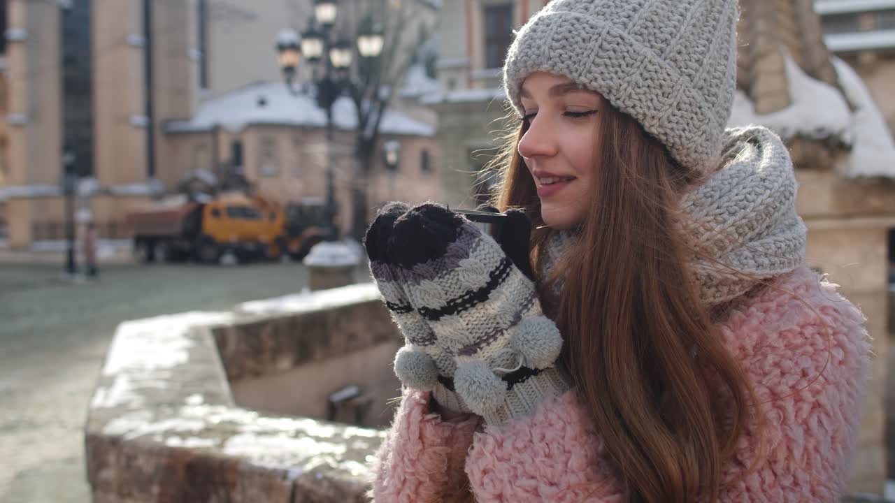 mujer joven con un sombrero, bufanda y guantes, sonríe mientras bebe una taza de café al aire libre en invierno