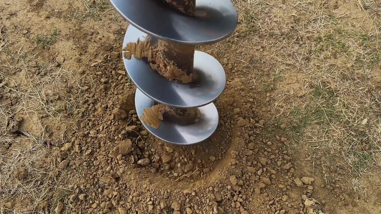 Vista ravvicinata di una punta da trapano a coclea che ruota nel terreno, creando un foro per un palo di recinzione, con terra smossa e ghiaia circostanti in un contesto di costruzione rurale all'aperto.