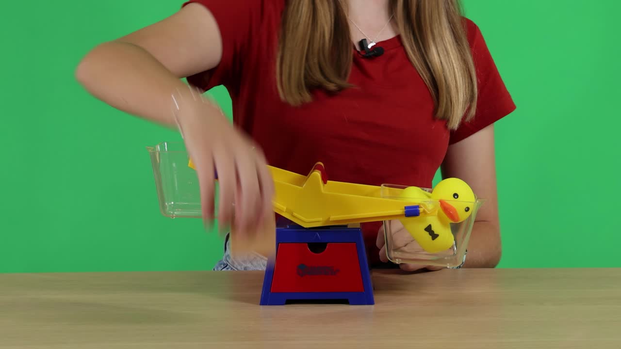 A person balances a toy duck on a colorful scale against a green background, demonstrating equilibrium principles