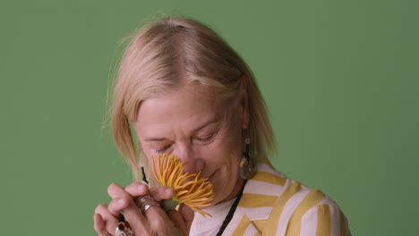 Blonde-Reife-Frau-Mit-Blauen-Augen-In-Gestreiftem-Hemd-Und-Accessoires,-Die-An-Einer-Sonnenblume-Riecht-Und-Auf-Grünem-Hintergrund-In-Die-Kamera-Lächelt-1