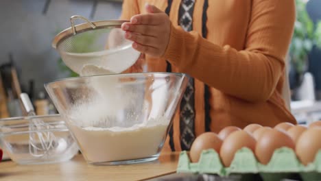 Video-of-midsection-of-biracial-woman-baking-in-kitchen-at-home,-sieving-flour-into-mixing-bowl