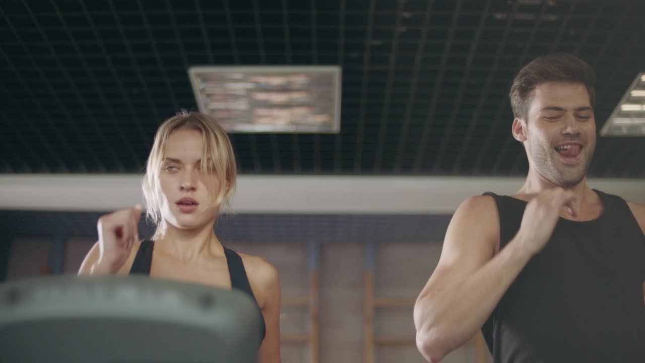 섹시한 스포츠맨이 러닝 머신에 있는 진지한 여자를 유혹하고 있습니다.