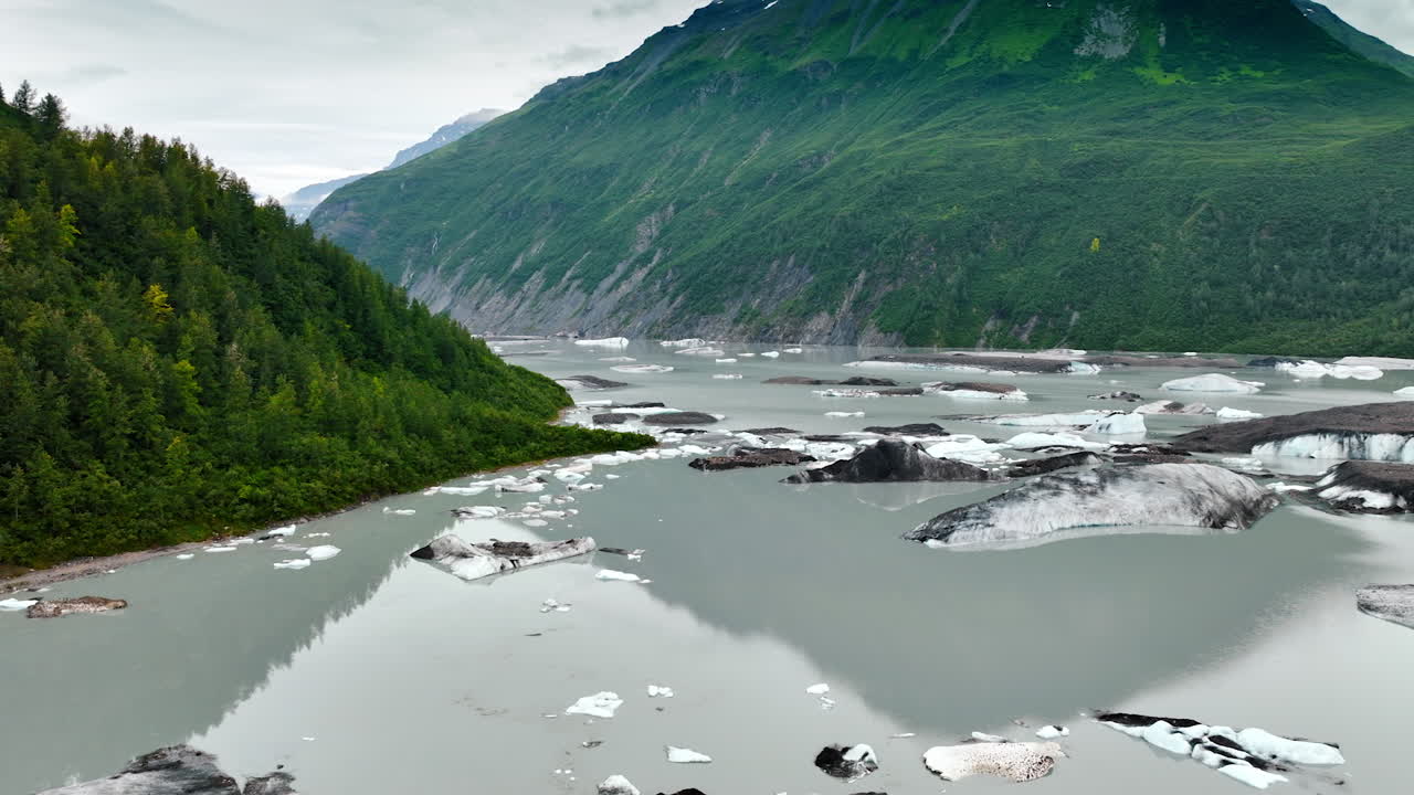 River flowing among the mountains colored grey with large pieces of melting ice. Rocks covered with evergreen forests at backdrop. Wild nature of Alaska