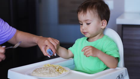 pretty caucasian boy eating from fork which he's holding in left hand. mom puts the fork into son's right hand and helps him to take some food.
