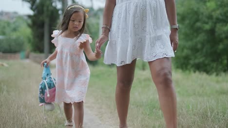 a pregnant woman and her young daughter, both dressed in white, walk hand-in-hand through a grassy park. the scene captures a serene moment of family bonding in nature, surrounded by trees.