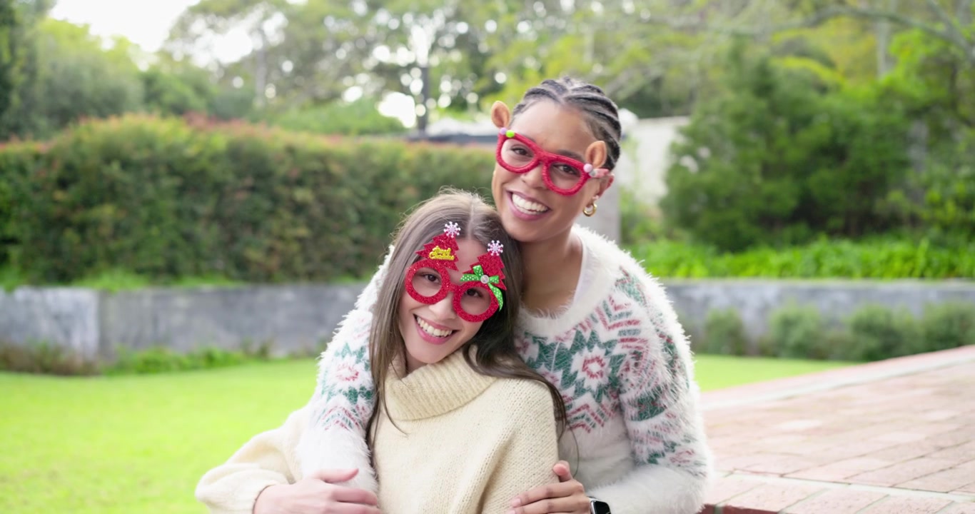 Two women 20s and 30s laughing and leaning on brick ledge in yard adjusting Christmas glasses