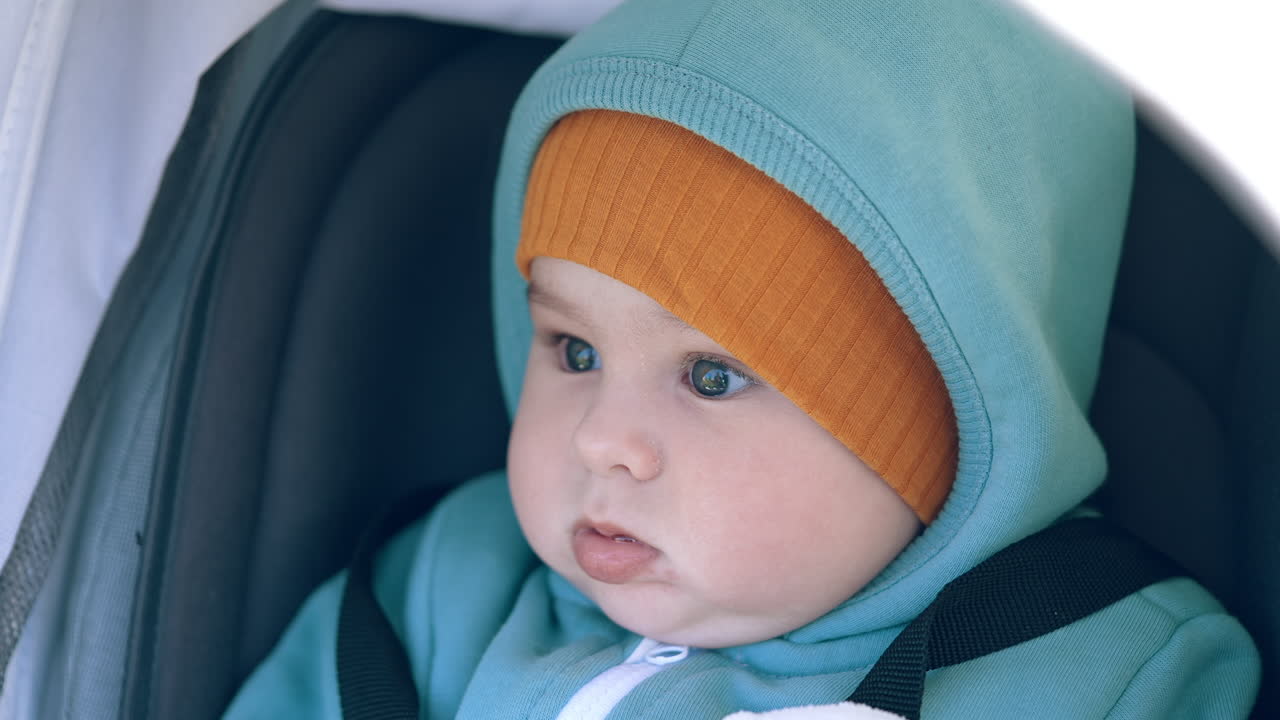 Lovely Caucasian boy with sweet cheeks sits in stroller. Cute child wearing warm clothes and cap having a walk in pram. Close up.