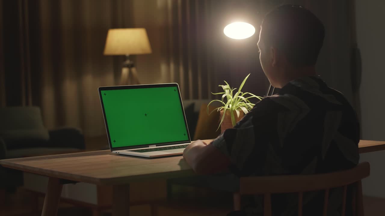 Teenage Boy Sitting At His Desk Learning Online On A Laptop With Green Mock-Up Screen. Late At Night In His Living Room Boy Uses Notebook Computer And Writing On Book. Over Shoulder Close Up View