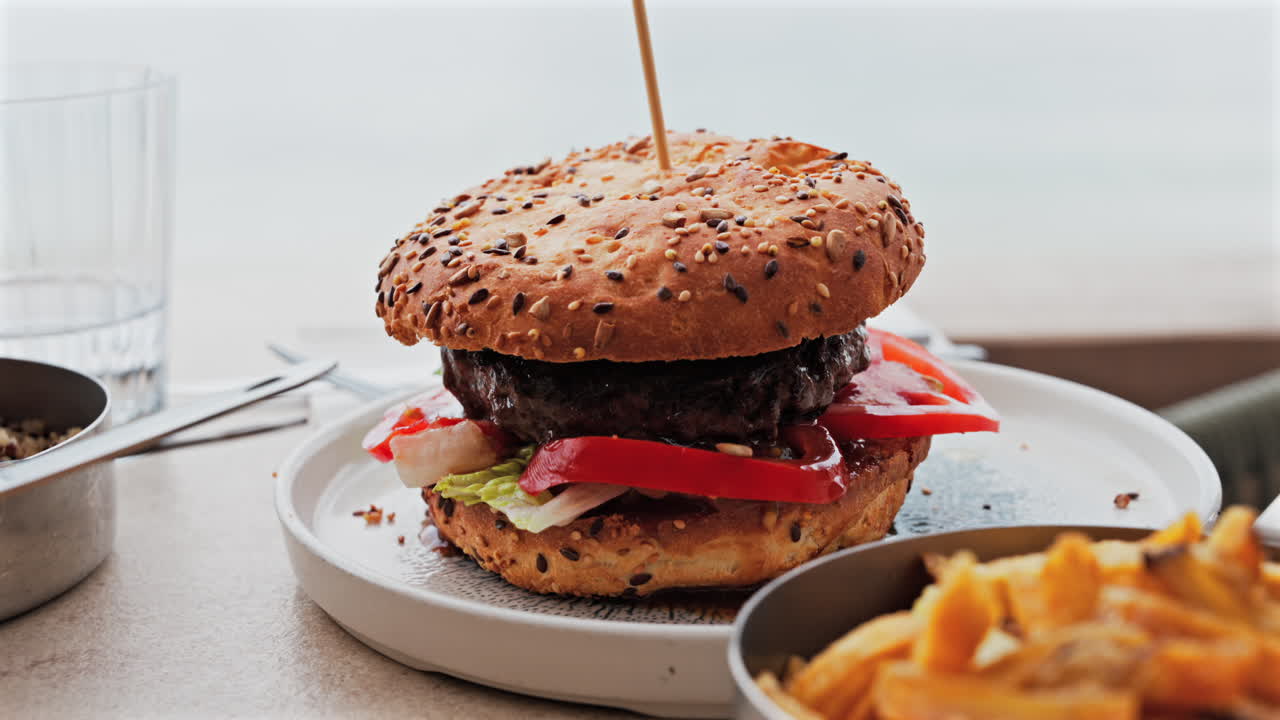 Close up of a hamburger on a white plate near french fries on a table at a restaurant