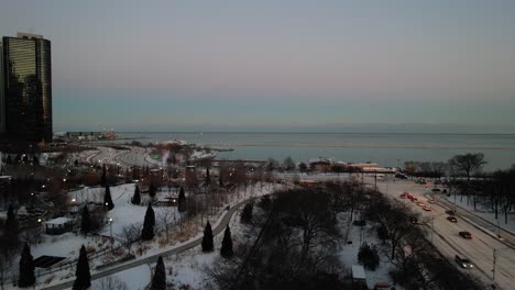 Aerial-shot-of-a-snowy-city-a-road-with-cars-driving,-a-park-with-trees-and-on-the-horizon-the-sea-in-Chicago