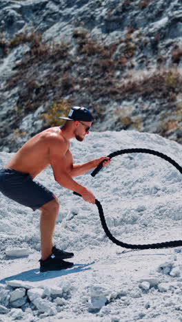 side view on bodybuilder training with battling ropes outdoors. handsome athlete quickly moves his hands with cables on the mountain background. vertical video