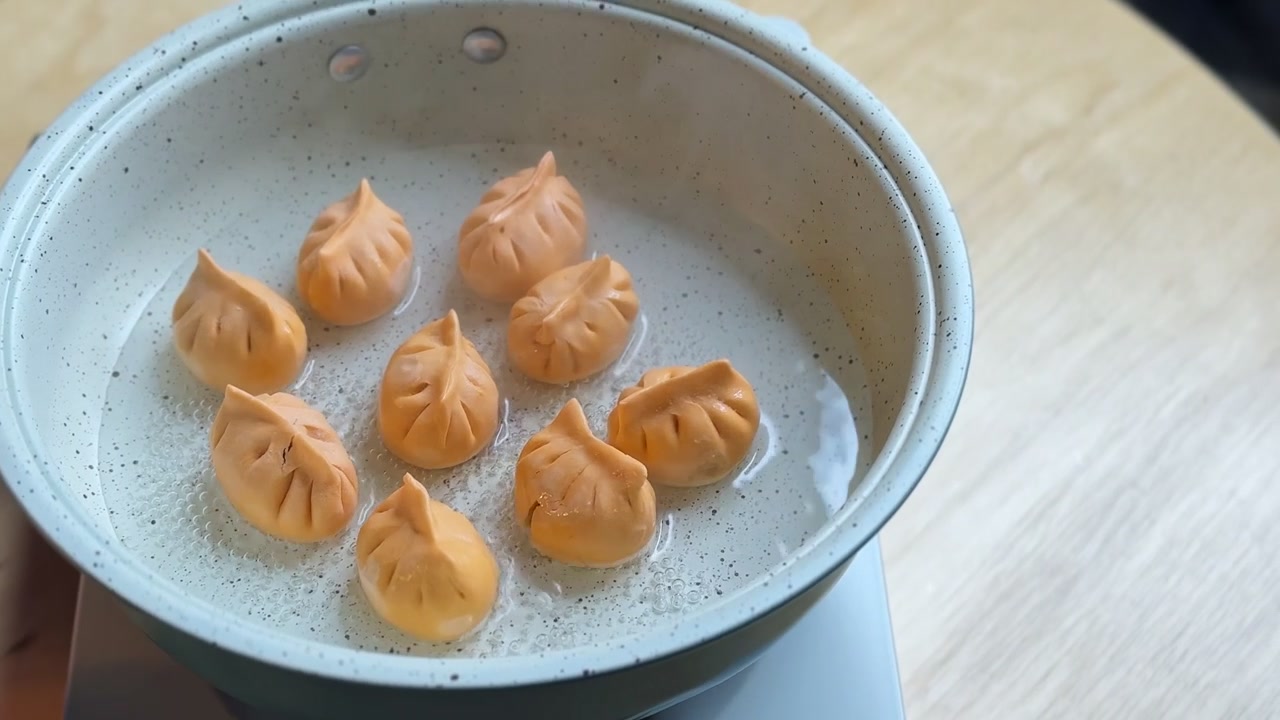 Uma mão coloca cuidadosamente bolinhos laranja em uma panela de água fervente em um fogão. Close-up em ângulo alto de dim sum asiático sendo cozinhado em um ambiente de cozinha moderna
