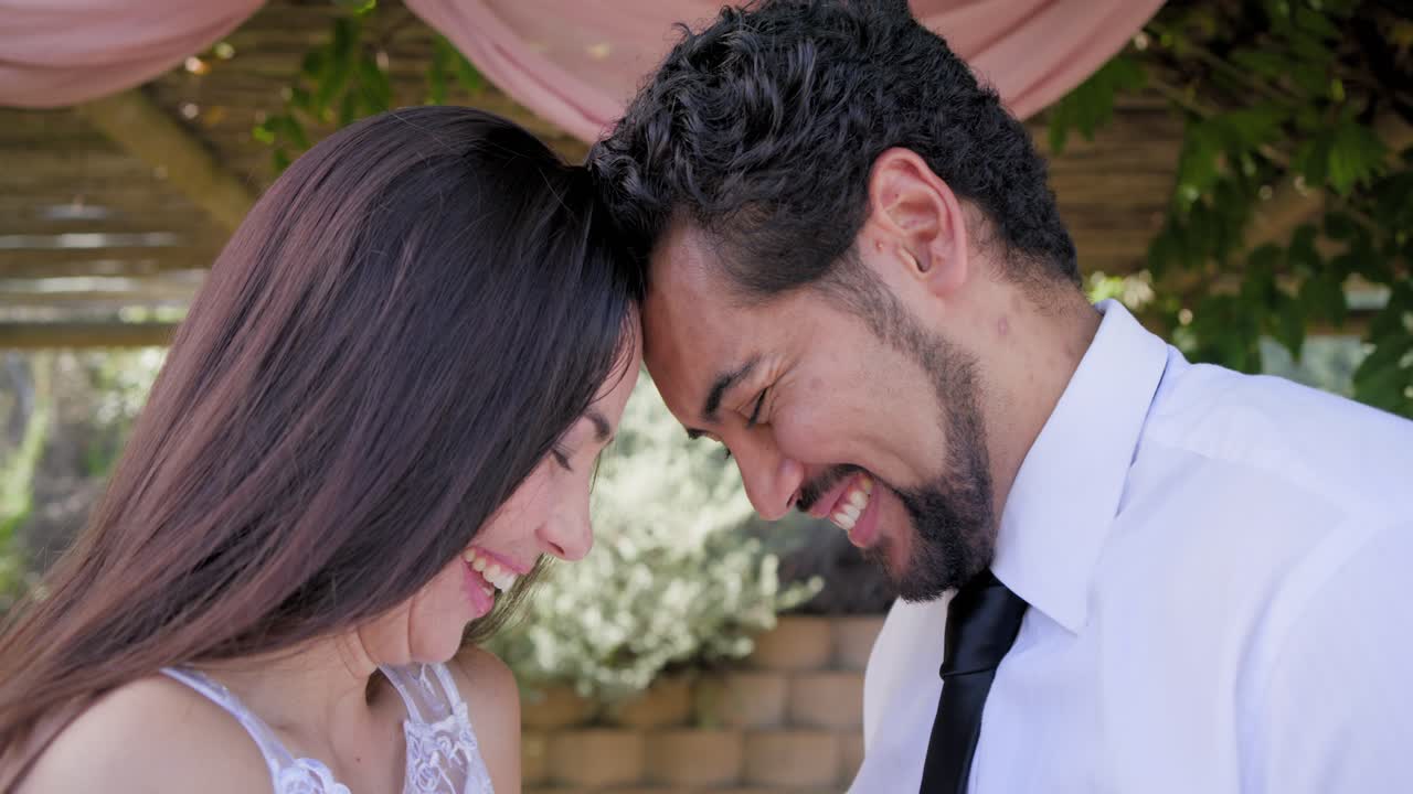 Pareja romántica bajo una pérgola con cortinas rosas, tocándose las cabezas, lo que provoca vítores de los invitados, y una mujer de encaje.