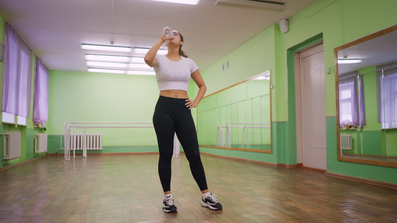 Young woman in white and black top standing indoors drinking water from clear bottle with calm relaxed posture after workout under natural light studio background highlighting hydration recovery