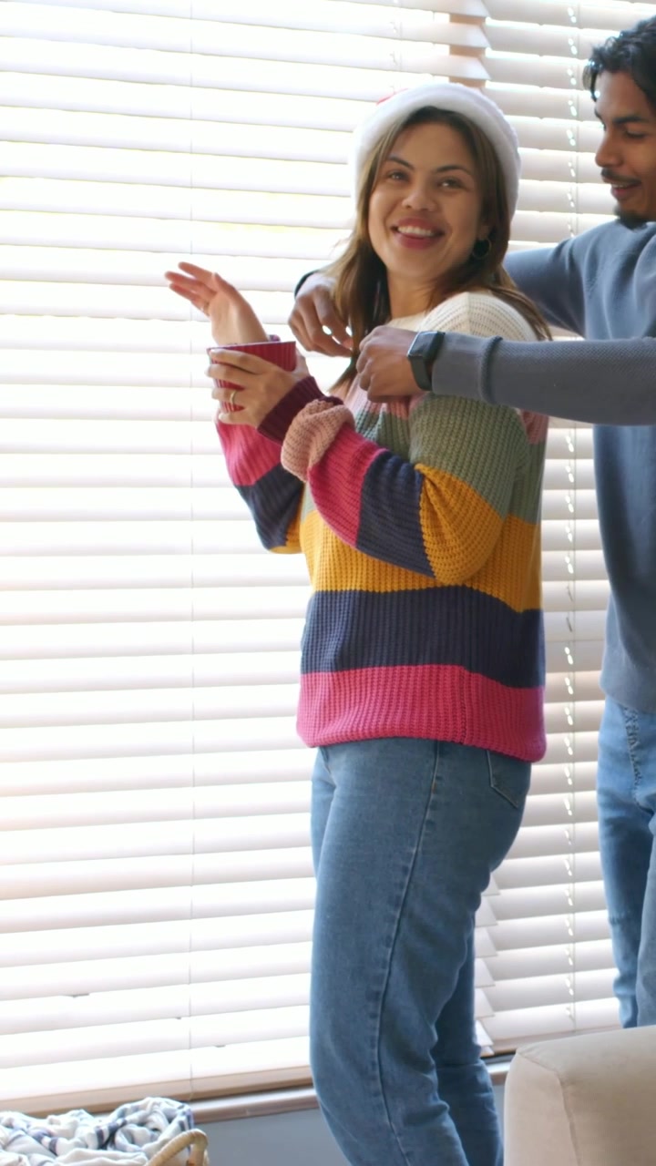 Video verticale: Donna con cappello di Babbo Natale che sbircia dalle persiane con una tazza, un uomo che arriva e l'abbraccia come saluto.