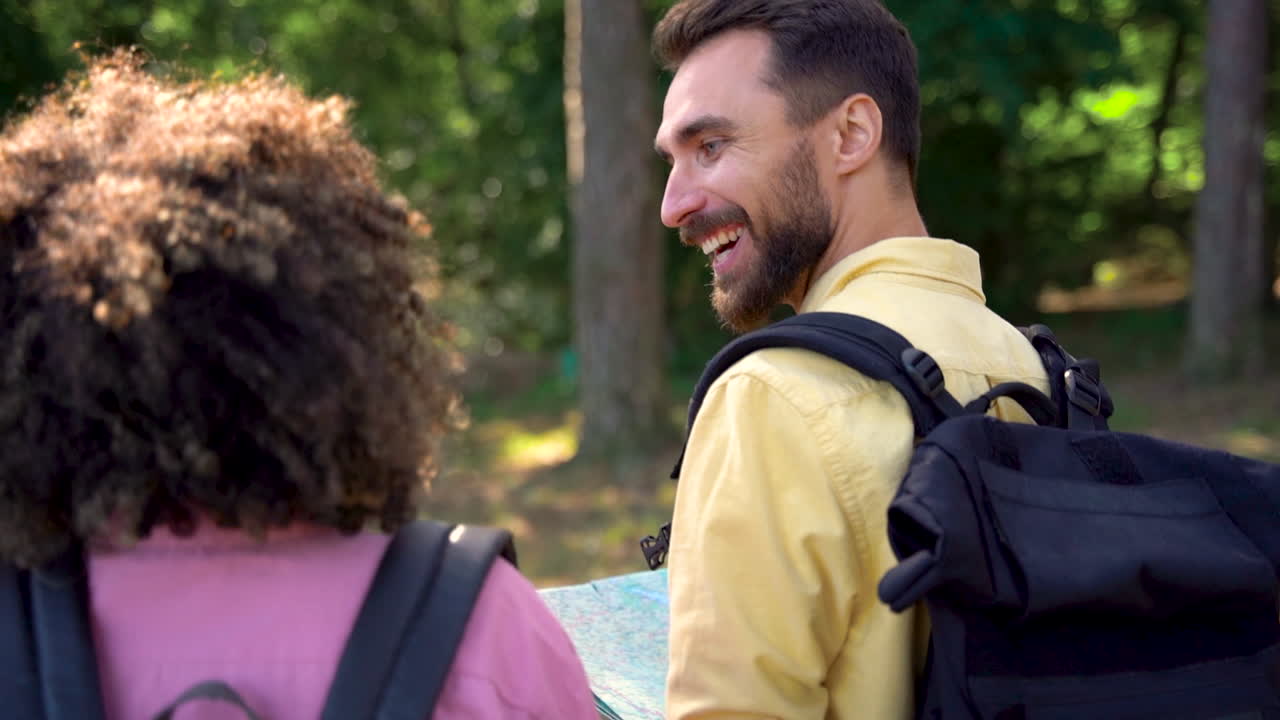 Hiker couple with backpacks and map walk and talk on a forest trail. View from behind. Young black female and caucasian man holding hands.