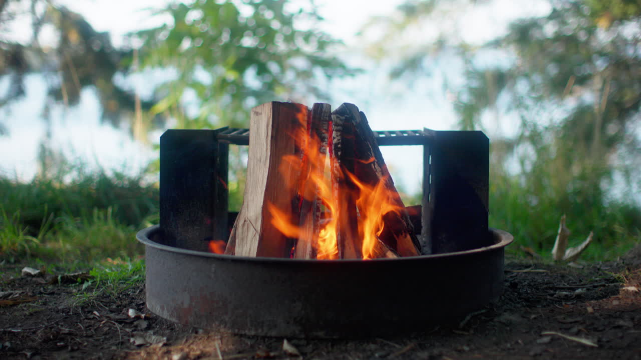 Handheld shot of a cozy campfire burning in a firepit, framed by lush greenery with the St. Lawrence River in the background at Wellesley Island State Park in Fineview, New York.