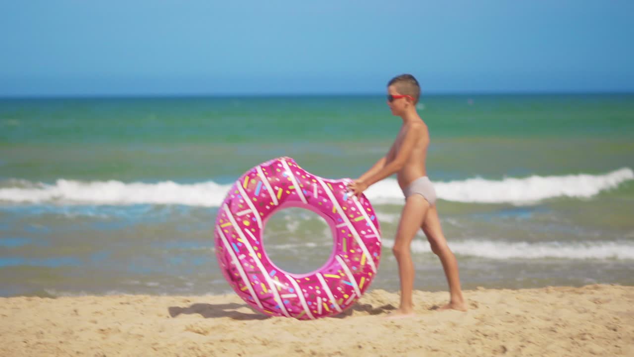 el niño corre a lo largo de la playa con una rosquilla inflable rosada, la enrolla a lo longo de la arena contra el fondo del mar. el concepto de relajación y diversión