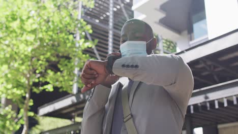 African-american-man-in-city-wearing-face,-using-smartphone-and-looking-at-smartwatch