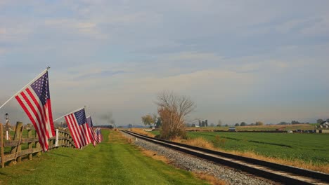 A-View-of-a-Single-Rail-Road-Track,-With-a-Steam-Passenger-Train-Approaching,-With-a-Fence-with-America-Flag-on-it,-Gently-Waving-in-the-Wind-on-a-Sunny-Autumn-Day