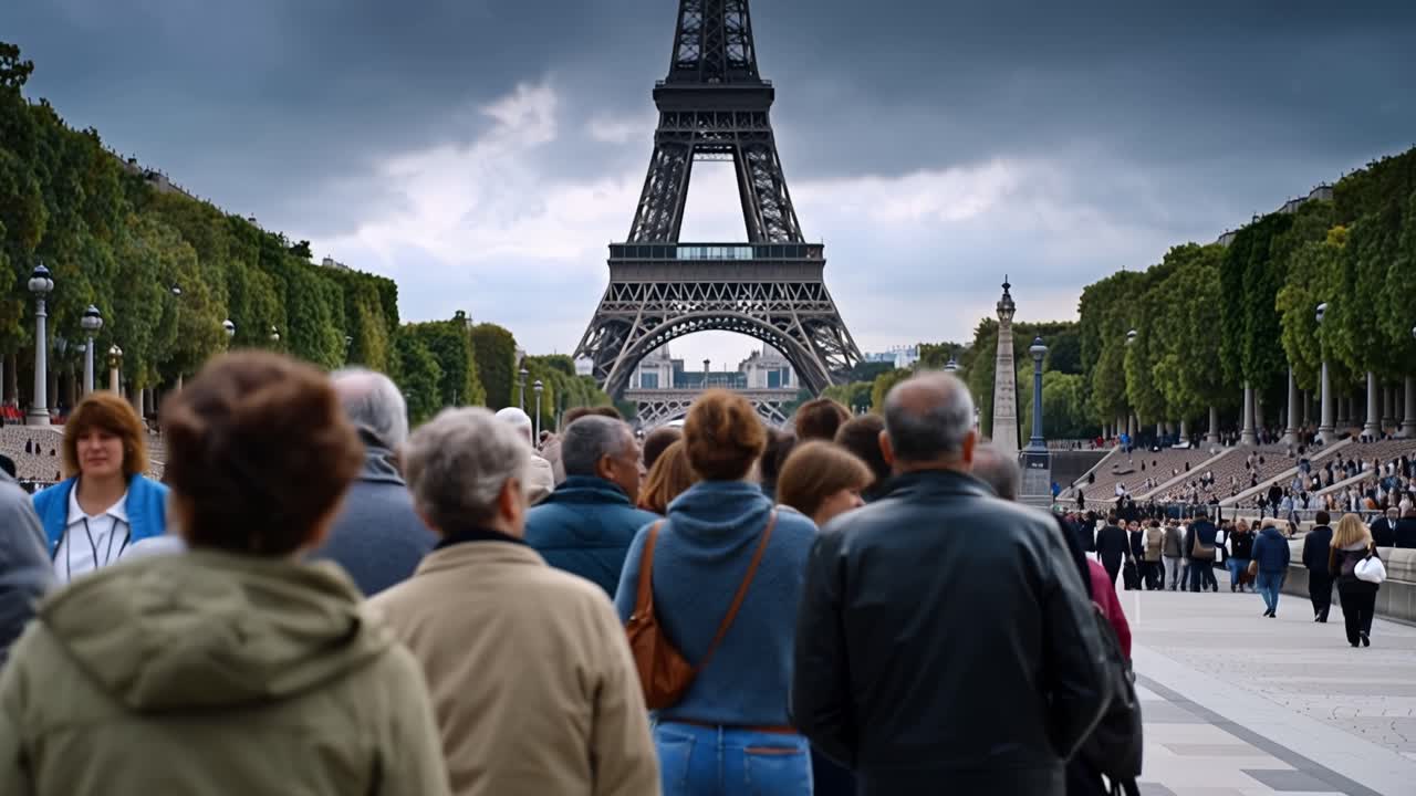 Folle di persone camminano verso la Torre Eiffel sotto cieli nuvolosi. Le persone appaiono calme, concentrate sul punto di riferimento davanti a loro. La scena cattura il turismo parigino, la vita cittadina e l'attrazione culturale.
