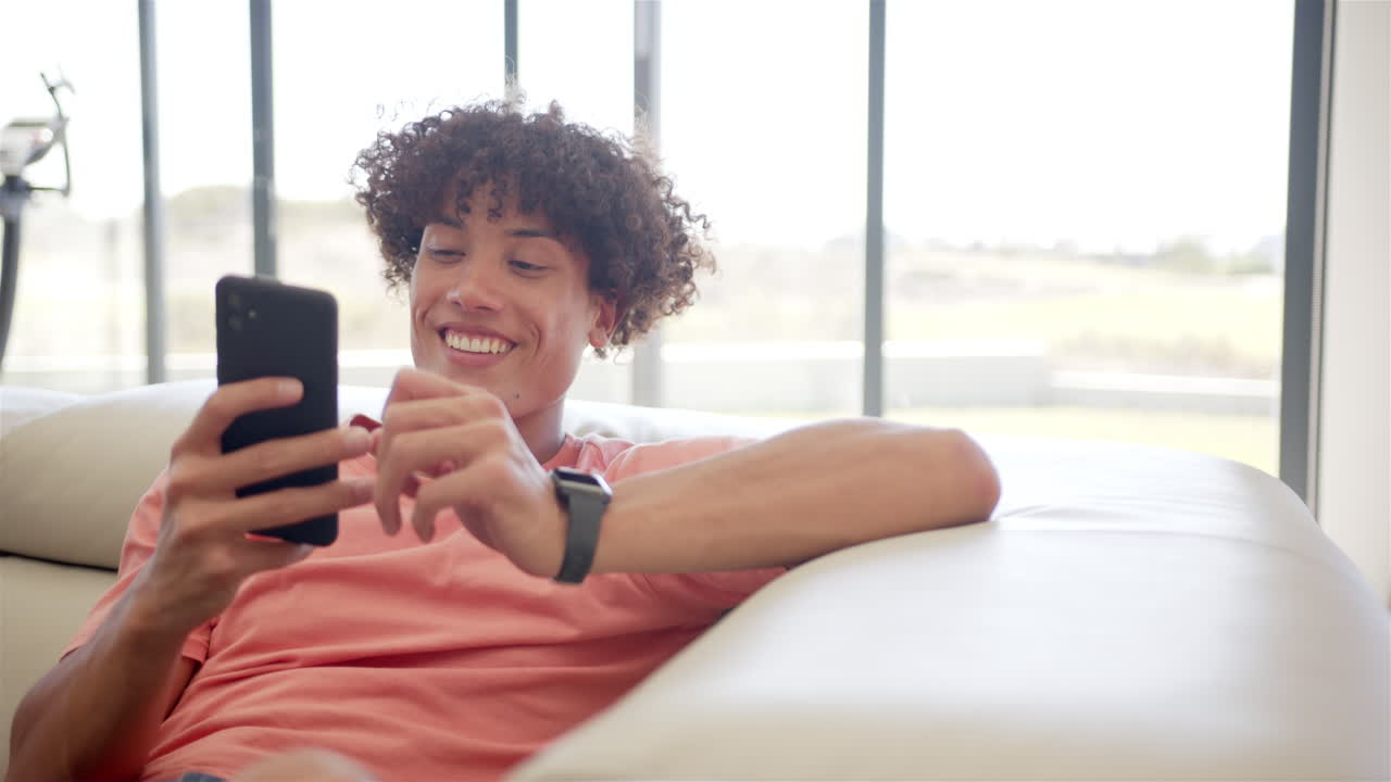 A young biracial man is engrossed in his smartphone while relaxing on a couch at home