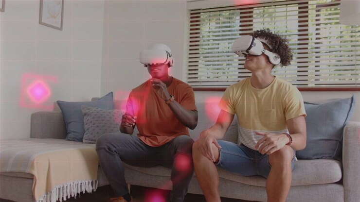 Due uomini su un divano, quello a sinistra alza la mano causando la comparsa di sfere rosa, indossano visori VR bianchi e tengono in mano i controller, giocando