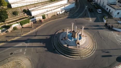 Toma-Aérea-Giratoria-Desde-Una-Calle-En-Medina-Sidonia-En-Cádiz-España-Con-Una-Rotonda-Con-Columnas-Y-Fuentes,-Casas-Blancas,-Tráfico-De-Automóviles-Y-Peatones-En-Un-Día-Soleado