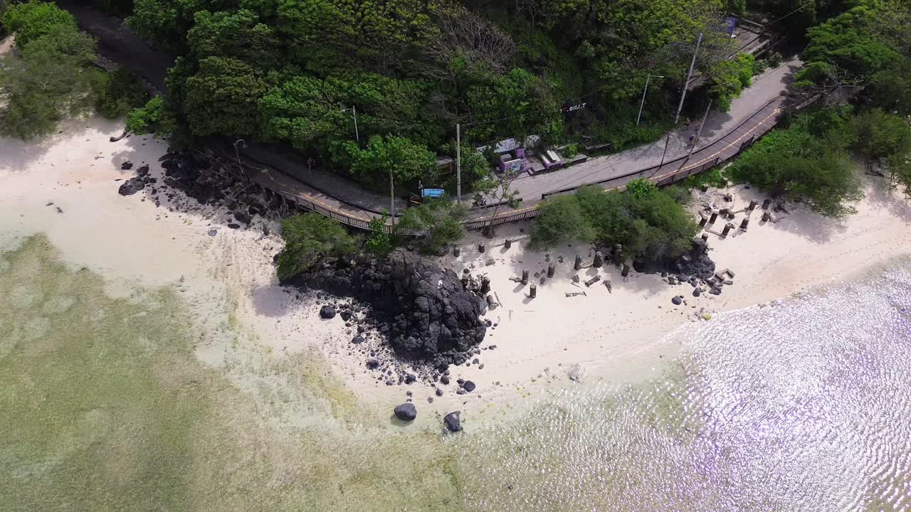 Top-down aerial static shot capturing white sand, black rocks, and tropical trees near road