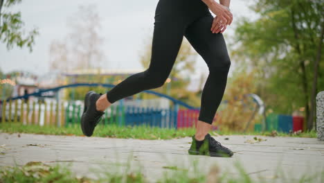 close-up view of a woman in black leggings and black sneakers stretching outdoors, background includes blurred lights, adding a park setting with leaves scattered on the ground