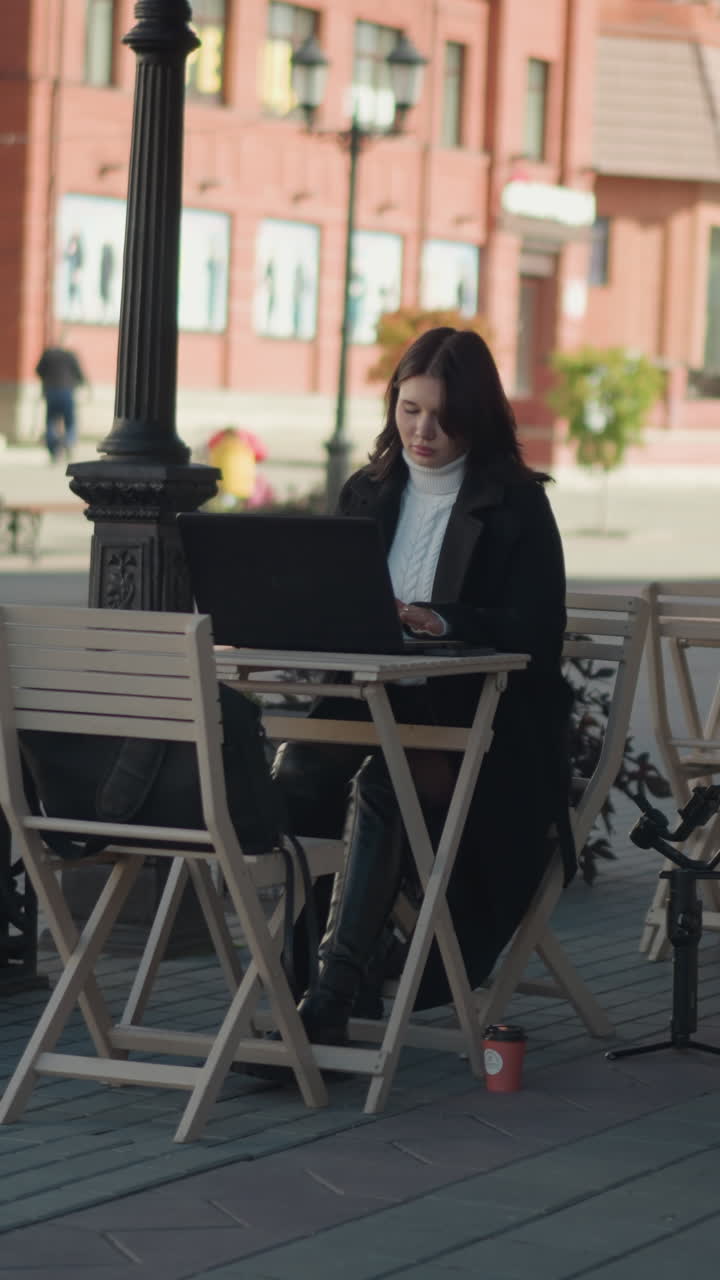 Woman with dark hair working on laptop at outdoor table in cozy cafeteria setting, surrounded by plants and chairs with blurred background showing city street, bus passing in the distant
