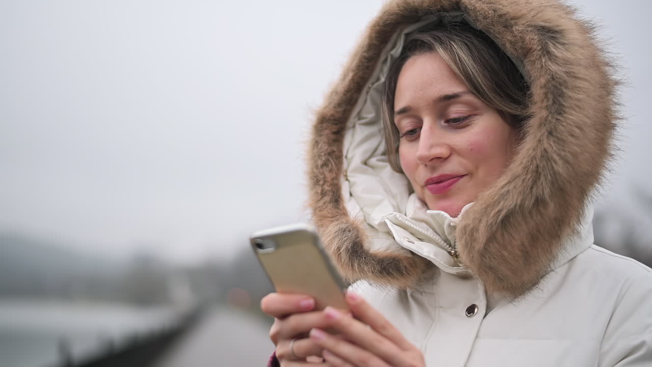 Женщина в теплом пальто с меховым капюшоном проверяет свой смартфон, стоя в парке. Погода облачная и прохладная, что создает безмятежную атмосферу.