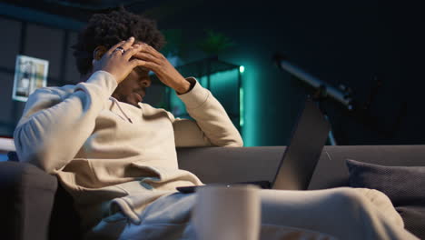 black person having health problems suffering from headache and nausea, sitting in the living room and struggling to work on tasks. remote worker suffering from illness at home. camera b.